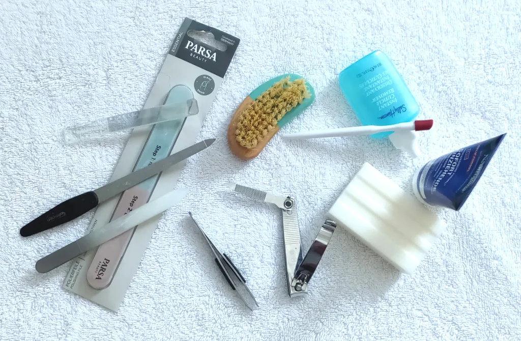 Hand care items on a white towel: glass file, sapphire file, glass buffer, three-step buffer, nail brush, cuticle hoof, cuticle remover, hand cream, white soap, nail clippers with cleaning tool, and tweezers.