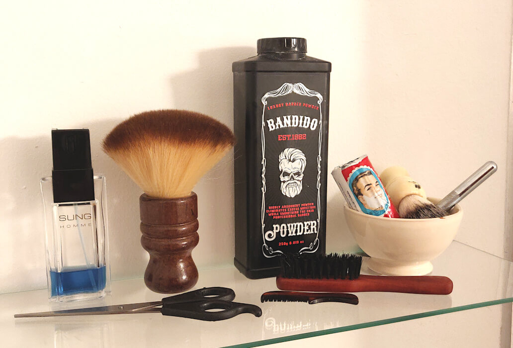 A curated display of classic grooming tools, including barber powder, a neck duster, a shaving kit in a ceramic bowl, a beard brush, a mustache comb, hairdressing scissors, and a bottle of cologne—evoking traditional barbershop rituals and refined masculine care.