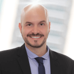 Professional headshot of a bald man in a suit. His scalp shows exaggerated bald head shine, suggesting healthy grooming.