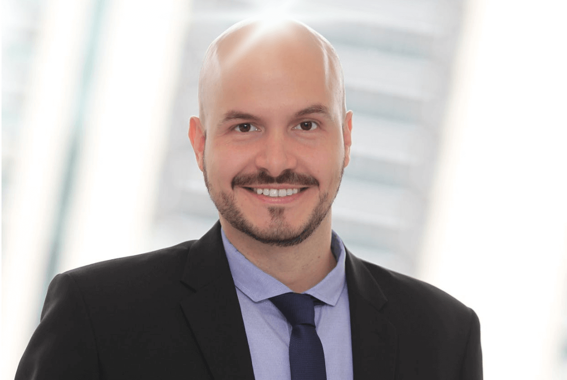Professional headshot of a bald man in a suit. His scalp shows exaggerated bald head shine, suggesting healthy grooming.