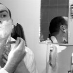 A man shaving in front of a mirror with traditional tools—part of a morning routine that may rely on grooming substitutes when essentials are missing.