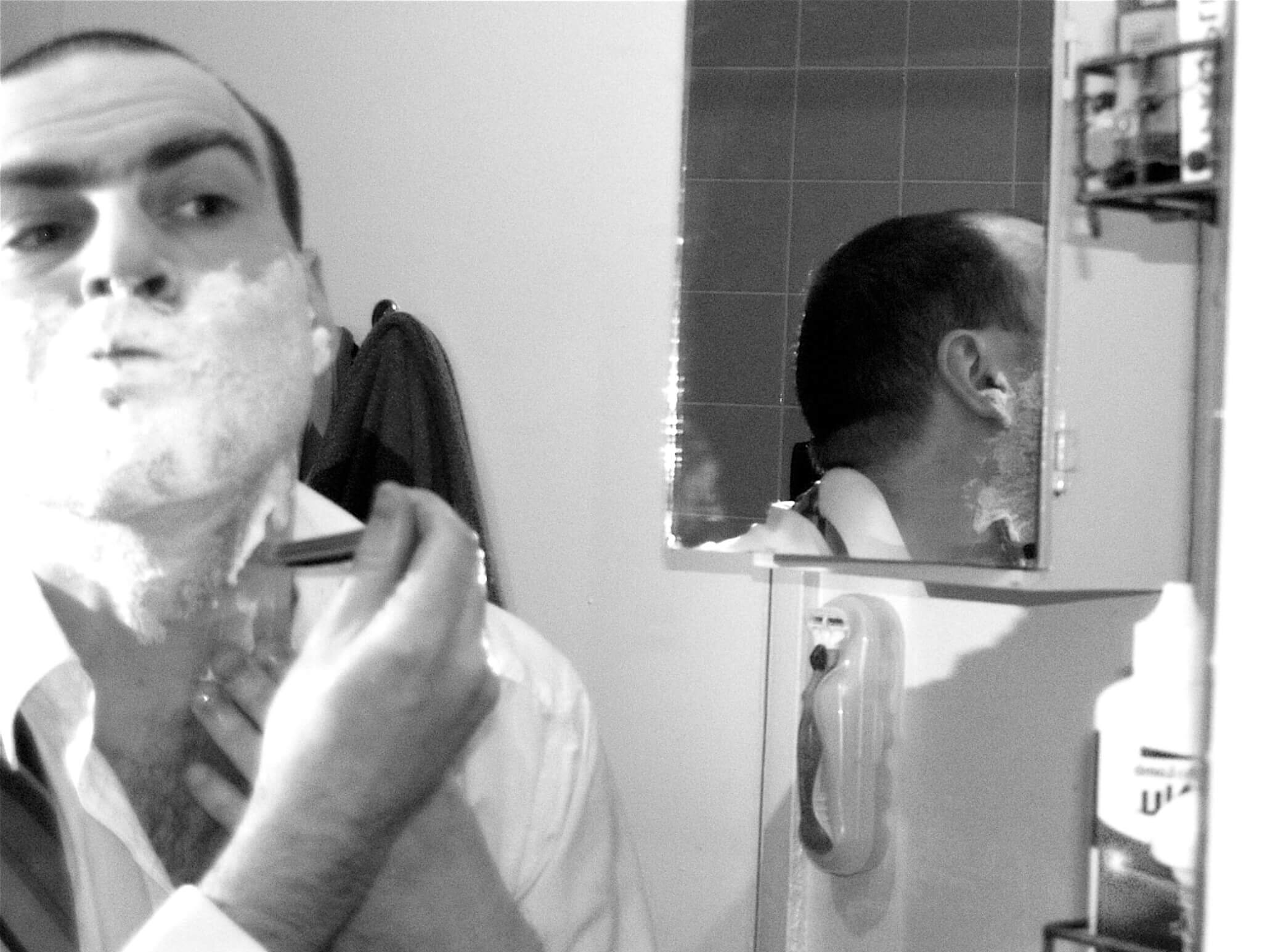 A man shaving in front of a mirror with traditional tools—part of a morning routine that may rely on grooming substitutes when essentials are missing.