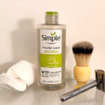 Flat lay of men’s grooming essentials showing a bottle of Simple micellar water, cotton pads, a shaving brush, and a safety razor arranged on a glass surface.