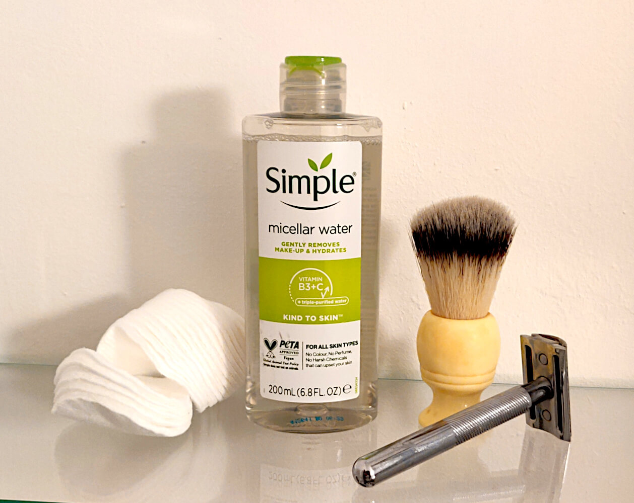 Flat lay of men’s grooming essentials showing a bottle of Simple micellar water, cotton pads, a shaving brush, and a safety razor arranged on a glass surface.