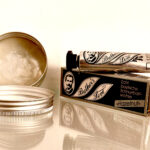 A tin of beard balm and two tubes of Brother's Love mustache wax, one in clear and one in hazelnut, sitting on a glass shelf.