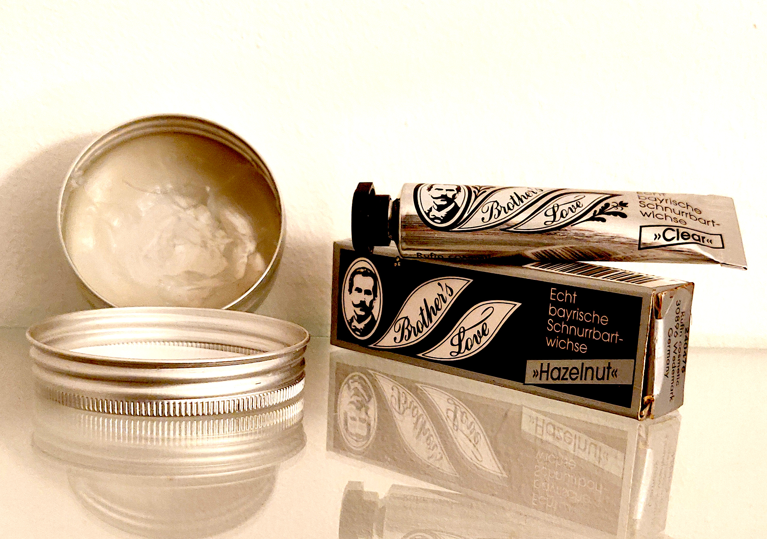 A tin of beard balm and two tubes of Brother's Love mustache wax, one in clear and one in hazelnut, sitting on a glass shelf.