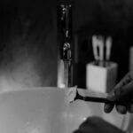 Hand rinsing a razor under running water while shaving the face, with shaving cream visible on the blade.
