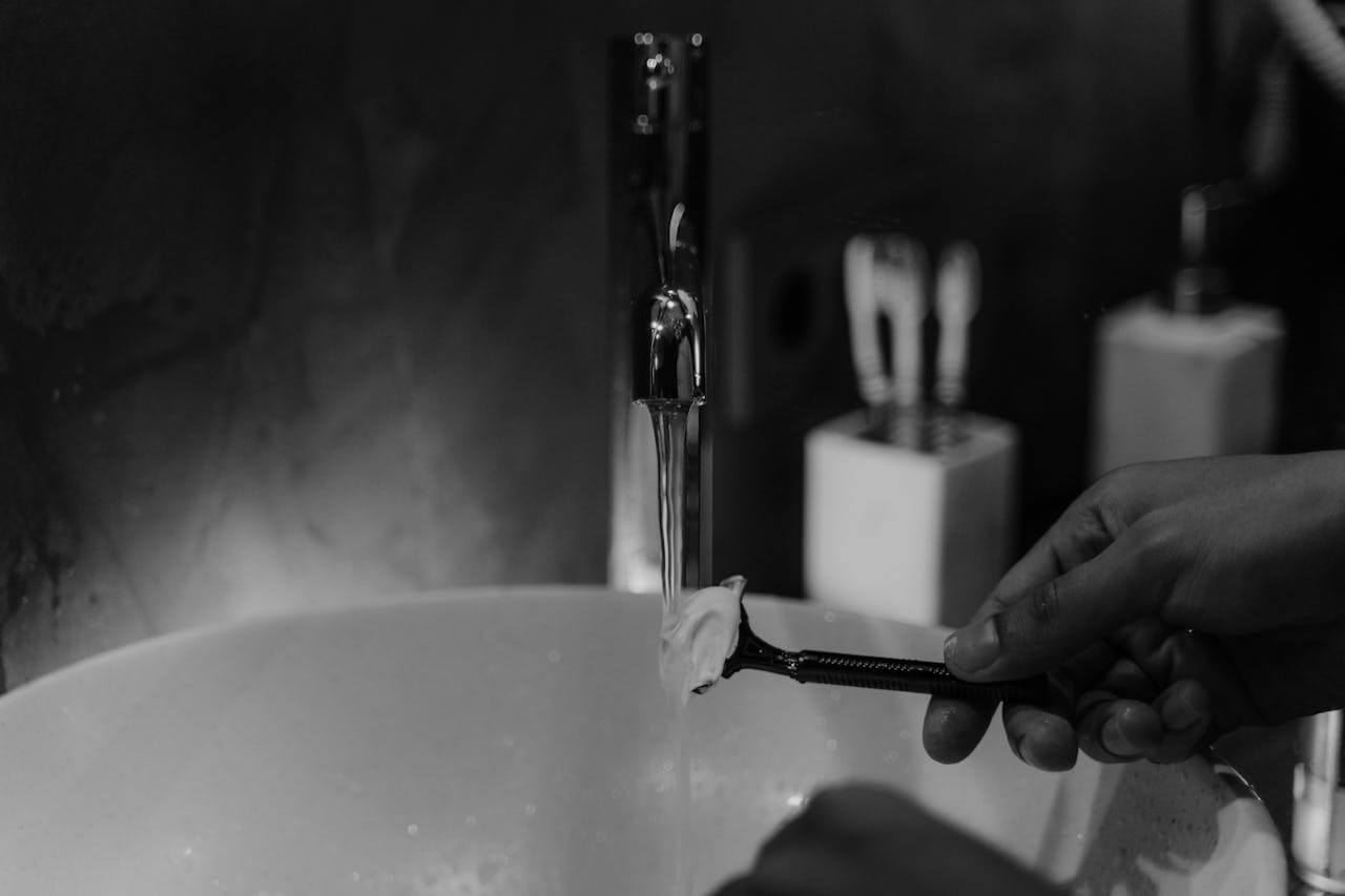 Hand rinsing a razor under running water while shaving the face, with shaving cream visible on the blade.