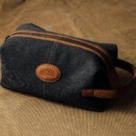 Dark gray fabric men’s toiletry bag with brown leather accents, a zipper, and a stitched oval patch reading ‘MAN CODE,’ placed on a brown textured surface.