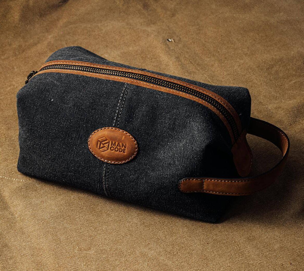 Dark gray fabric men’s toiletry bag with brown leather accents, a zipper, and a stitched oval patch reading ‘MAN CODE,’ placed on a brown textured surface.