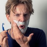 A man shaving with a disposable razor, reacting with discomfort as the blade drags across his skin—a realistic moment that reflects the irritation behind razor burn and razor bumps.