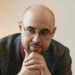 Man with glasses resting his chin on his hands, showing a bald head with a matte finish.
