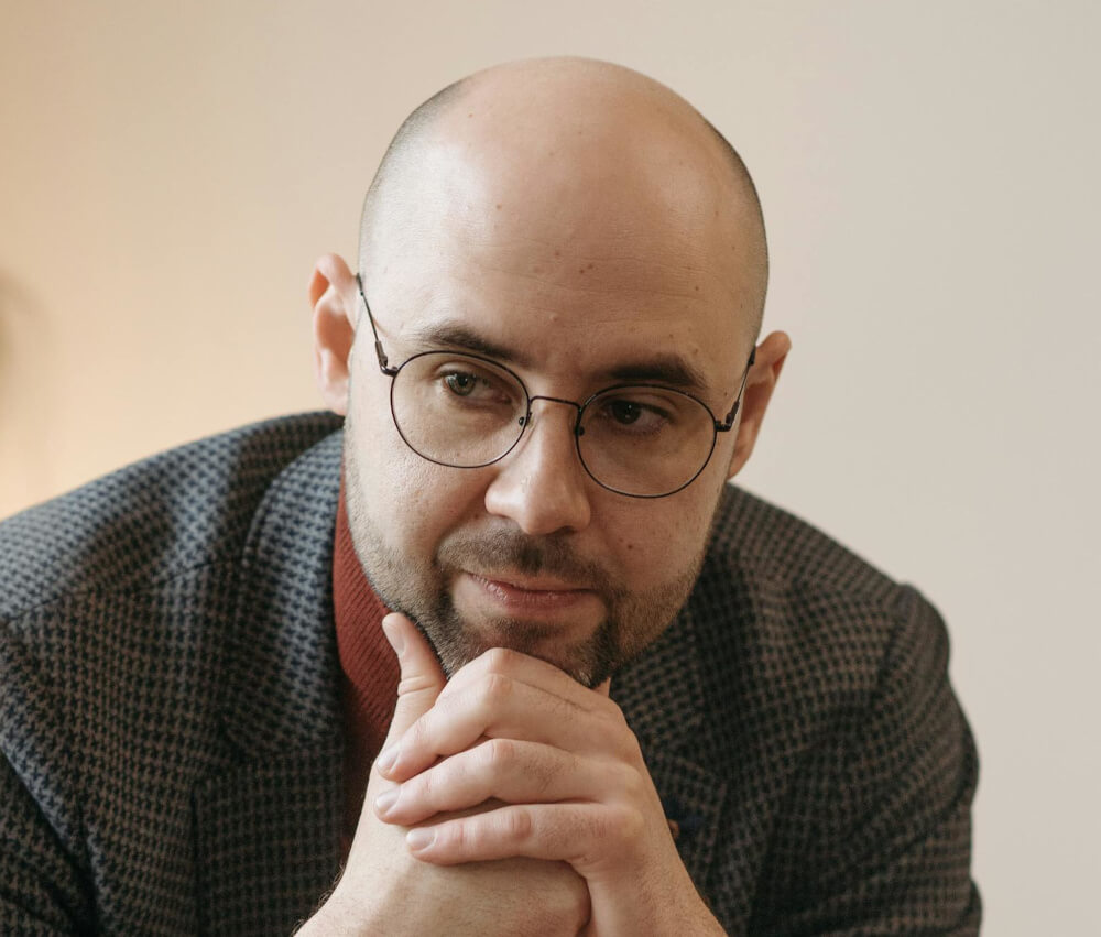 Man with glasses resting his chin on his hands, showing a bald head with a matte finish.