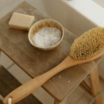 A wooden stool holding a bar of soap, a bowl of salts, and a long-handled bath brush beside a filled tub.