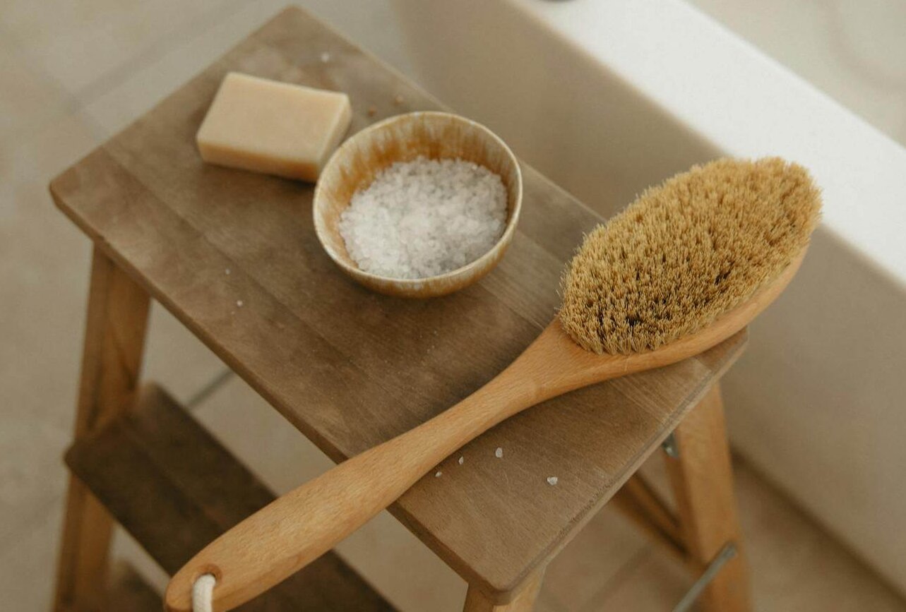 A wooden stool holding a bar of soap, a bowl of salts, and a long-handled bath brush beside a filled tub.