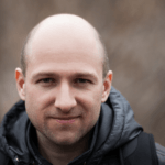 Close‑up portrait of a bald man with stubble wearing a dark hooded jacket outdoors, his horseshoe fringe visible.