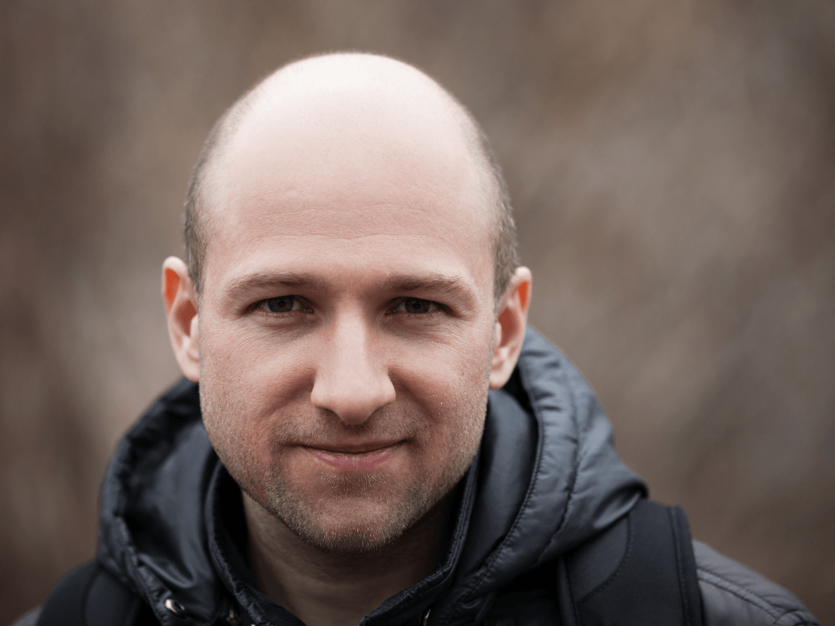 Close‑up portrait of a bald man with stubble wearing a dark hooded jacket outdoors, his horseshoe fringe visible.