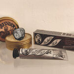 Open Defender tin with a guitar pick stuck into the balm, shown alongside Brother’s Love Bavarian mustache wax products to illustrate what men compare when choosing beard balm or mustache wax.