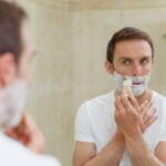 A man applying shaving cream before a bathroom mirror.