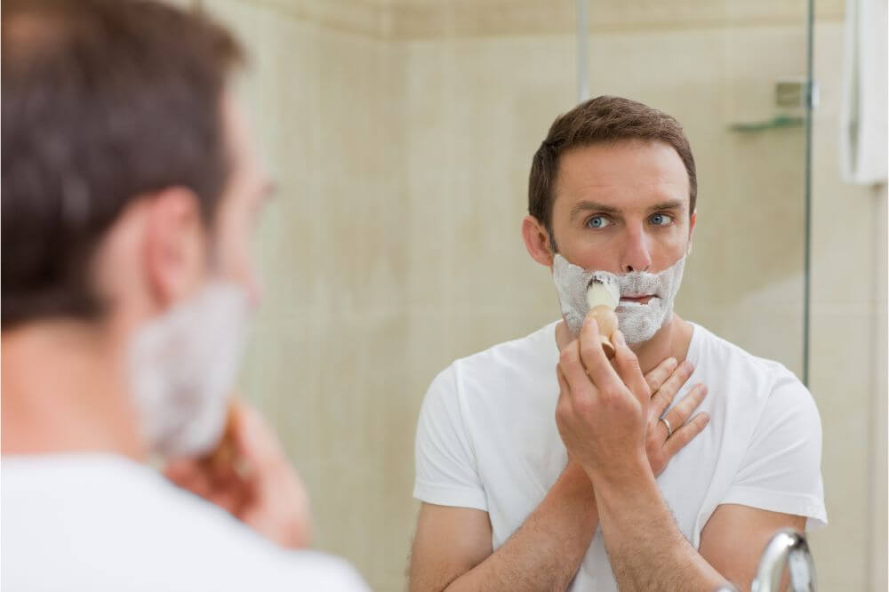 A man applying shaving cream before a bathroom mirror.