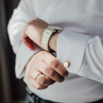 A man wearing discreet jewelry: a watch, a think wedding band, a mother-of-pearl cufflink.
