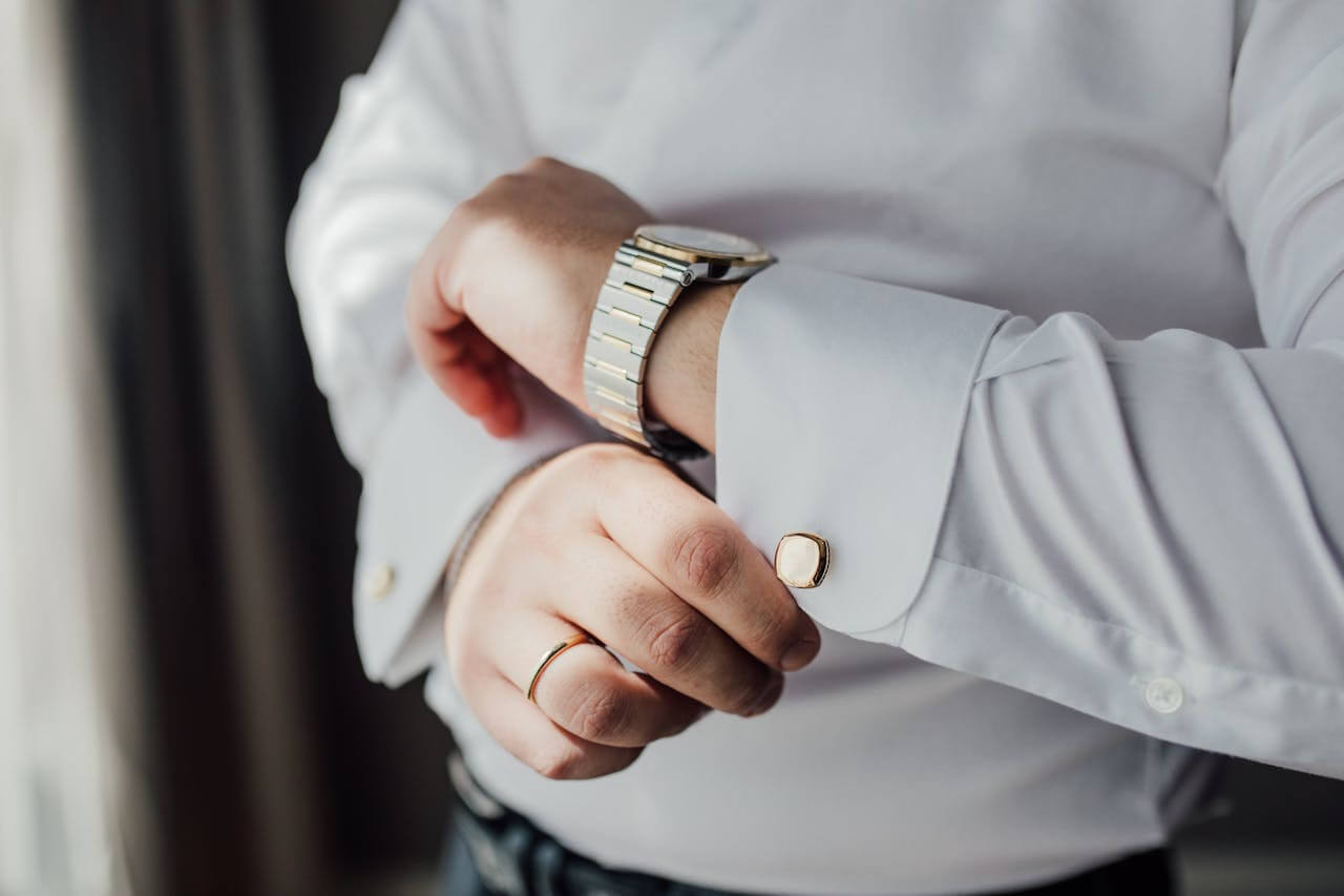 A man wearing discreet jewelry: a watch, a think wedding band, a mother-of-pearl cufflink.