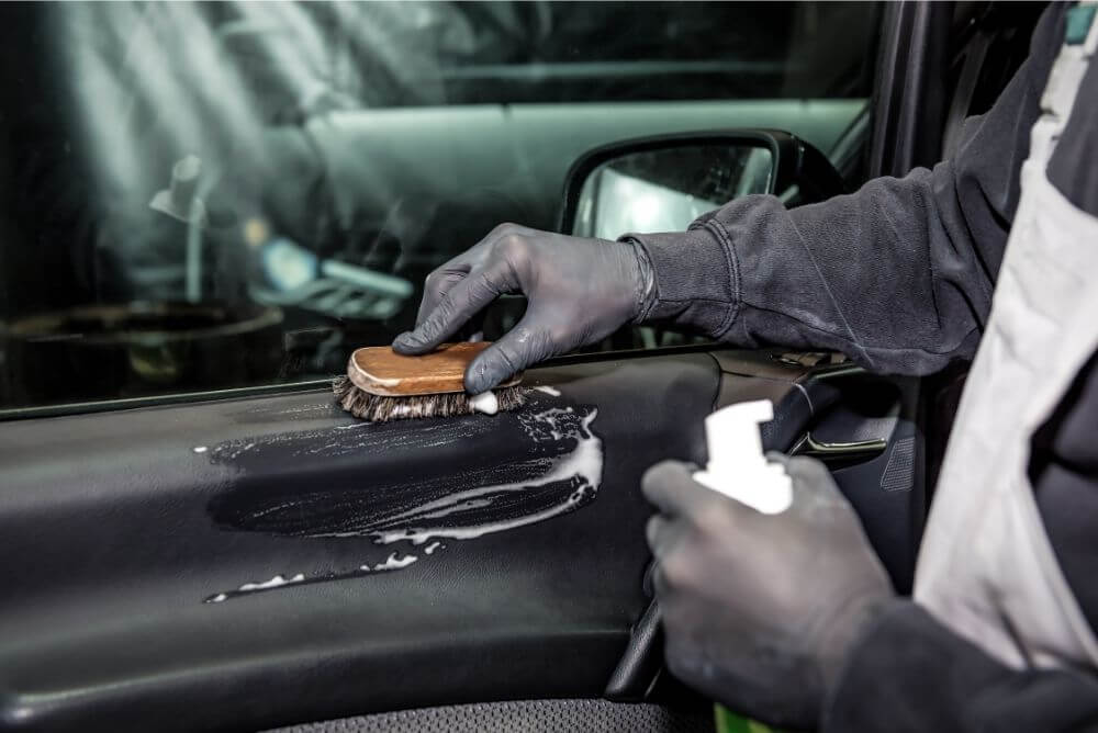 Gloved person scrubbing a car’s interior door panel with a detailing brush and cleaning solution.