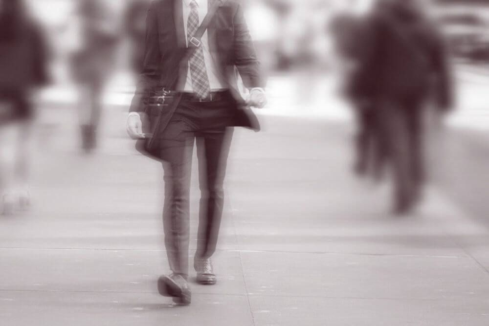 A man in a suit walking briskly along a city street, with motion blur suggesting a long exposure.