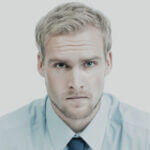 A pale blond man wearing a light blue shirt and dark blue tie.