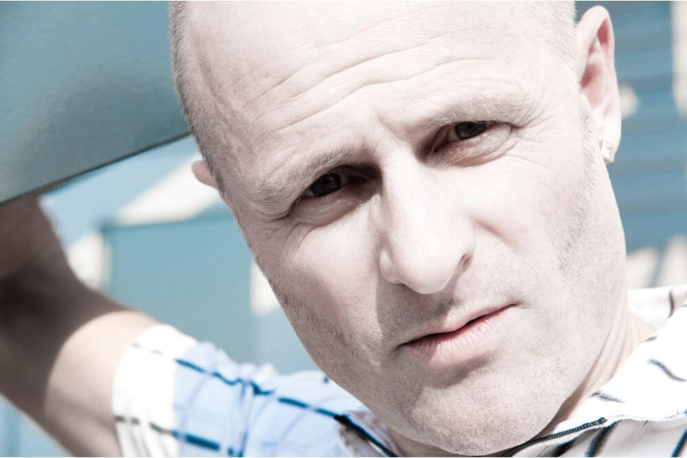 Bright outdoor close-up of a blond man with buzzed hair, showing a naturally pale skin tone.