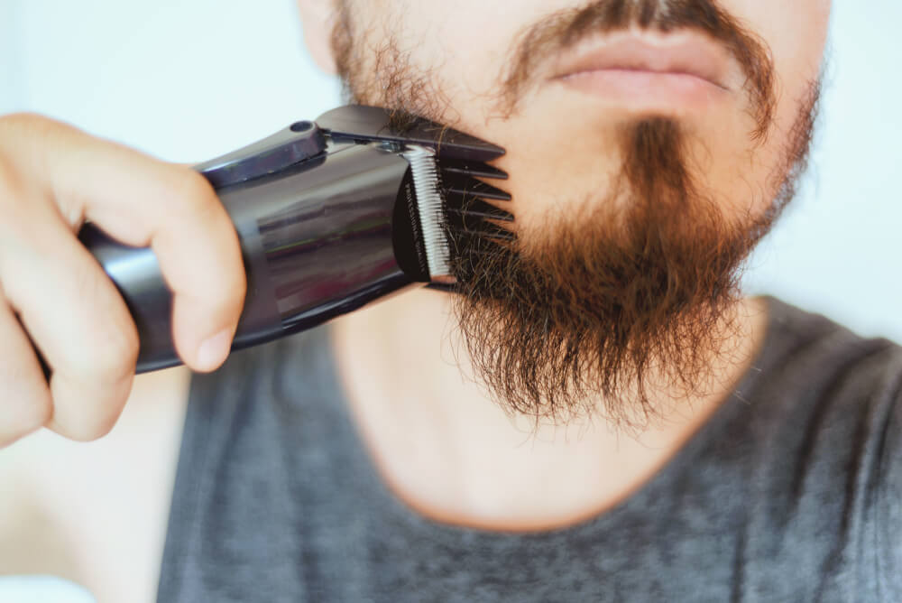 A man using a trimmer on his patchy beard.