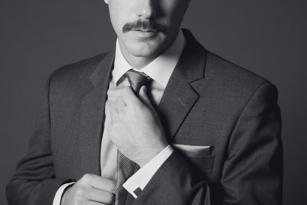 A man in a suit adjusting his tie, with cufflinks and a pocket square visible.